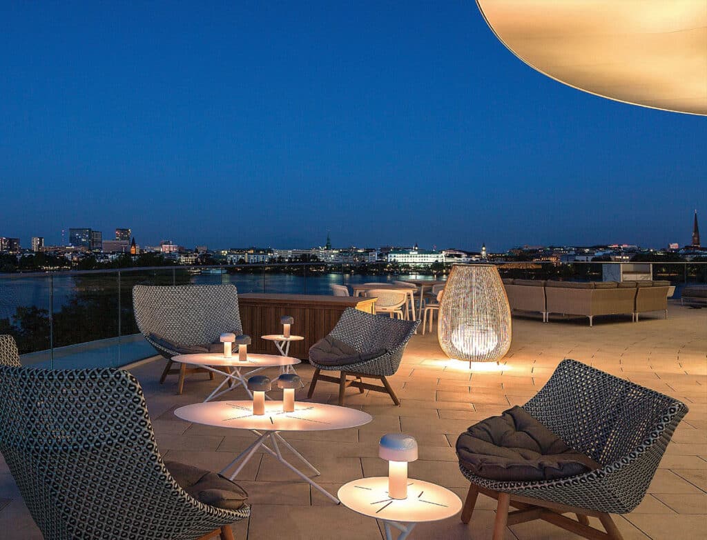 Die Bar-Dachterrasse des Hotels The Fontenay in der Abenddämmerung mit Körbstühlen in romantischer Beleuchtung. Im Hintergrund das abendlich erleuchtete Alsterpanorama.